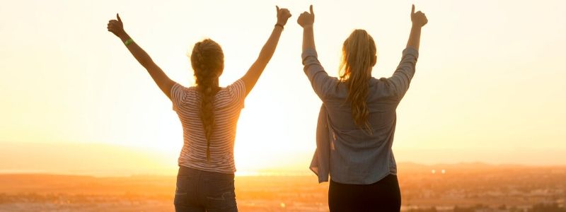 girls with raised hands looking over the city from a hill during sunset
