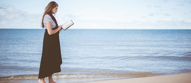 Young woman walking along the beach reading the bible near the water