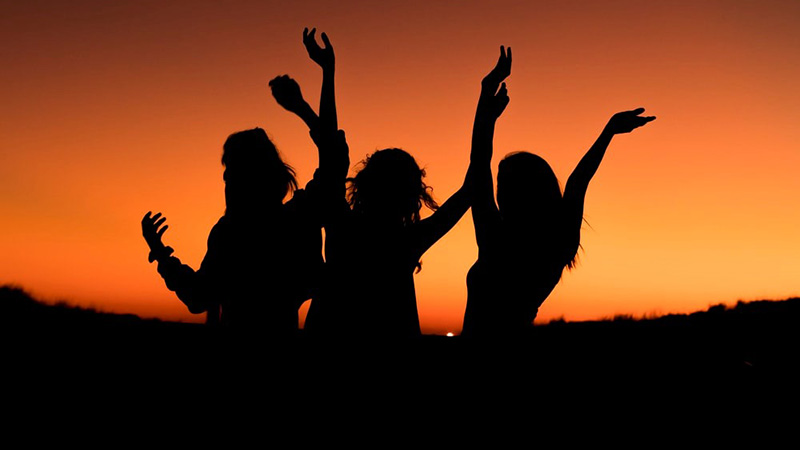 Black figures of three girls holding up their hands in the orange light of a setting sun