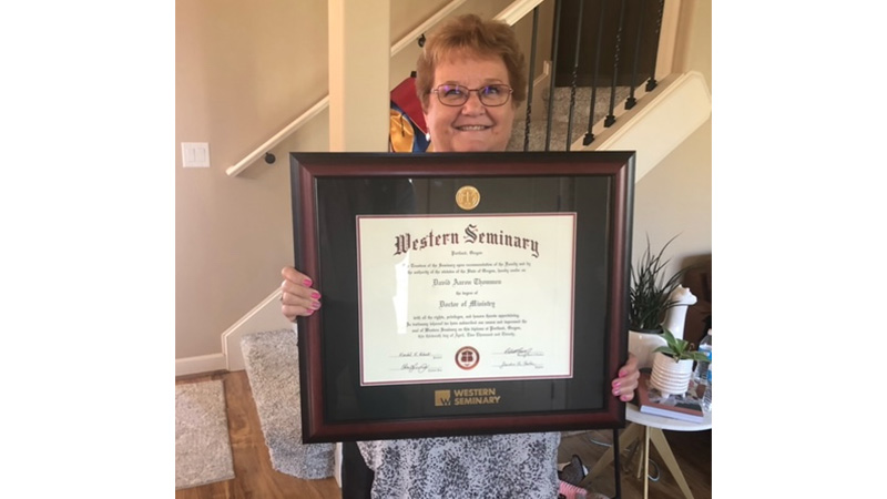 Glenda smiling in the foyer while holding her son's western seminary degree in a beautiful black and brown frame